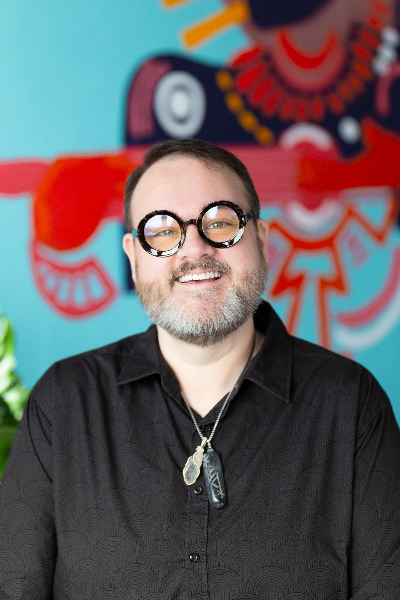 A profile image of a person smiling with glasses, a black shirt, a necklace on, and a painting behind them.
