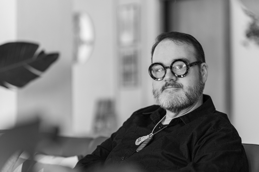 A black and white image of a person with glasses on and a necklace with a plant to the left and a building in the background.