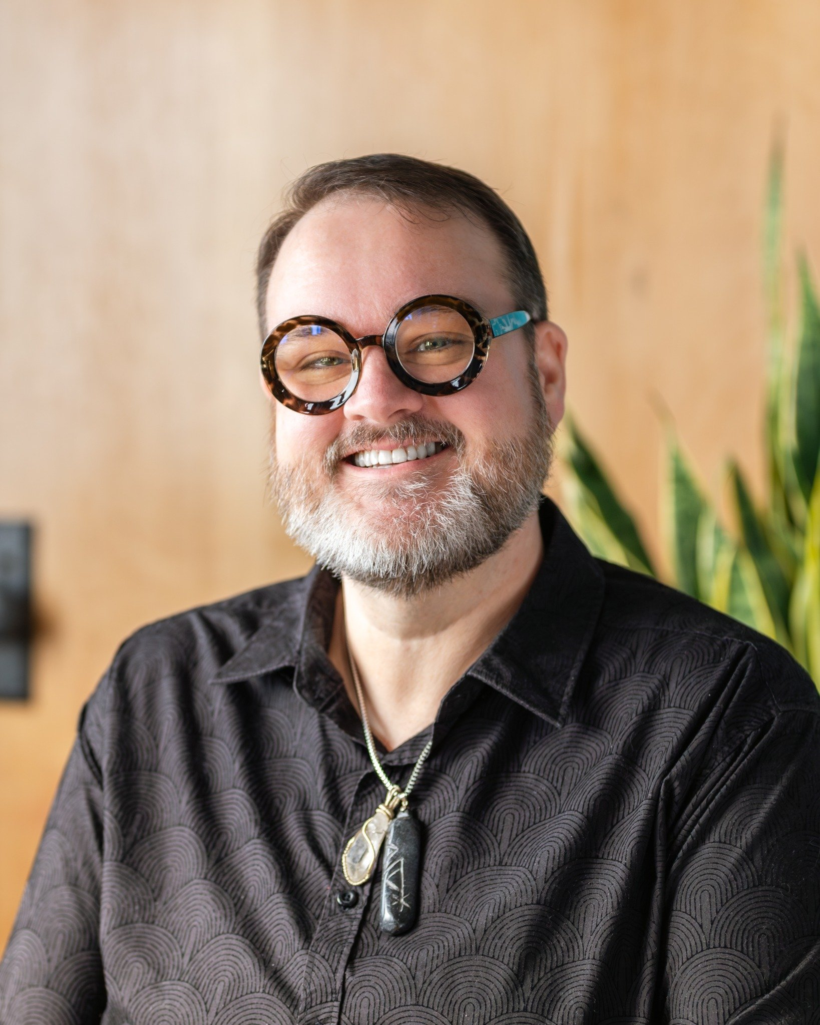 Image of profile image of a person with a black shirt, glasses, a necklace, and a plant behind them.